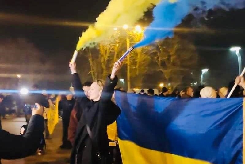 [ai] A crowd of people gathered at night, some holding a large blue and yellow flag, while a woman raises blue and yellow smoke flares. Several light sources illuminate the scene.